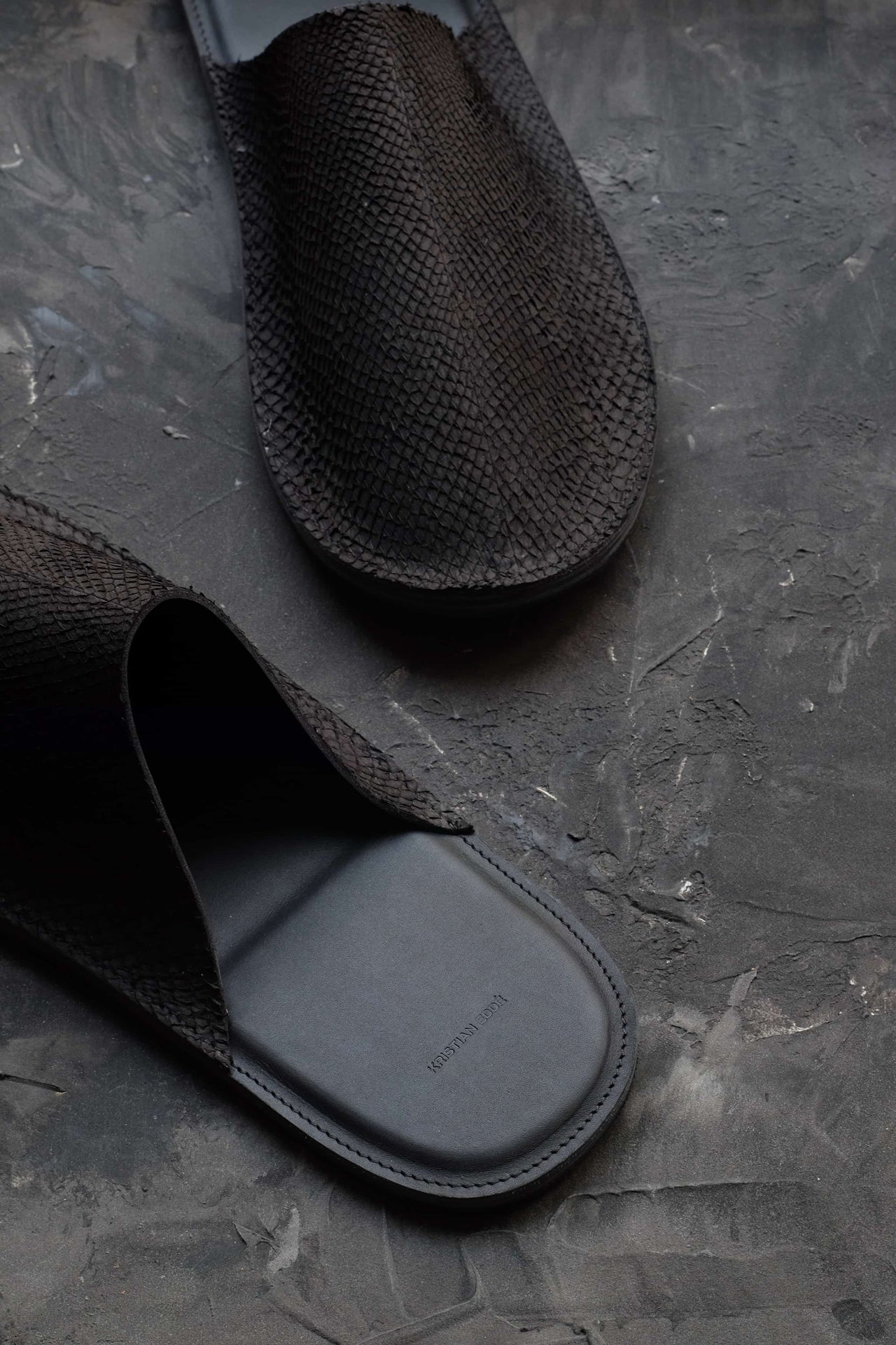 Detail of black salmon leather and embossed heel logo on handmade luxury slipper by Kristian Eddé